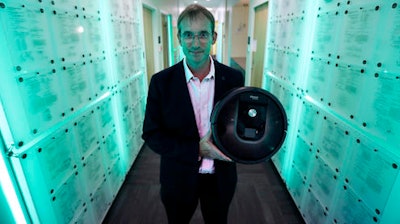 In this Thursday, Aug. 25, 2016, photo, iRobot co-founder and CEO Colin Angle is illuminated in blue-green light while holding a Roomba vacuum in a hallway decorated in patents the company owns, at their headquarters in Bedford, Mass. Angle is transitioning the company from military projects to household robots.
