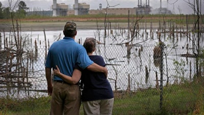 In this April 25, 2014, file photo, Bryant Gobble, left, embraces his wife, Sherry Gobble, right, as they look from their yard across an ash pond full of dead trees toward Duke Energy's Buck Steam Station in Dukeville, N.C. Duke Energy has agreed to remove millions of tons of coal ash containing toxic heavy metals from a power plant in North Carolina. The nation’s largest electricity company announced Wednesday, Oct. 5, 2016 that it would dig up three huge pits of water-logged ash at the Buck Steam Station near Salisbury. The ash will be dried and either offered for use in making concrete or moved to lined landfills elsewhere.