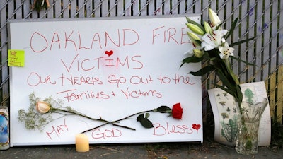 In this Dec. 5, 2016 file photo, signs and flowers adorn a fence near the site of a warehouse fire in Oakland, Calif. Attorneys representing the families of people who died in the Northern California warehouse fire that broke out during an unlicensed concert plan to file an updated lawsuit against the building's owner and manager Tuesday, May 16, 2017.