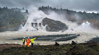 In this Feb. 20, 2017, file photo, workers and a firefighter watch water pouring down Oroville Dam's damaged main spillway in Oroville, Calif. Historical records reveal the late California Gov. Pat Brown misled voters about the cost of building the nation's tallest dam, ignored recommendations to delay construction and dismissed allegations that substandard materials were used to build the Oroville Dam.
