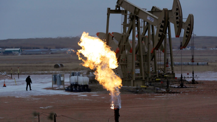 In this Oct. 22, 2015, file photo, workers tend to oil pump jacks behind a natural gas flare near Watford City, N.D. Fearing sanctions by the state, some North Dakota oil drillers have begun cutting output to control the amount of natural gas that's being burned off at well sites and wasted as a byproduct of crude production, industry and state officials say.