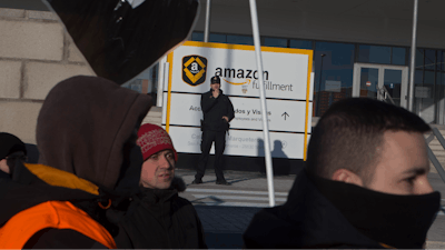A police officer stands in front of Amazon workers who gathered to protest outside the main Amazon Logistics Center where goods are stored for distribution, on the outskirts of Madrid, Spain, Wednesday March 21, 2018. Amazon workers are staging a 2 day strike over a labor dispute.