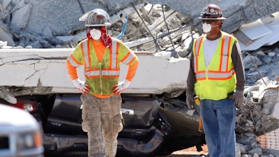 Workers stand in front of crushed cars under a section of a collapsed pedestrian bridge, Friday, March 16, 2018 near Florida International University in the Miami area. The new pedestrian bridge that was under construction collapsed onto a busy Miami highway Thursday afternoon, crushing vehicles beneath massive slabs of concrete and steel, killing and injuring several people, authorities said.