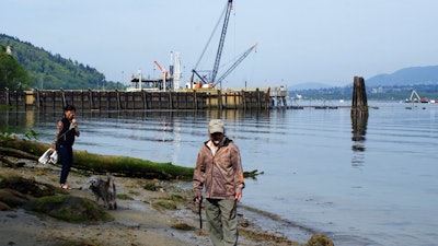 In this May 3, 2018 photo, a couple walk their dog on the shore near the Kinder Morgan Inc. Westridge oil tanker terminal in Burnaby, British Columbia, Canada. The company's Trans Mountain expansion is projected to increase oil tanker traffic here from about 60 vessels per year to more than 400, as the pipeline flow increases from 300,000 to 890,000 barrels per day.