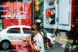 In this Oct. 9, 2014 file photo, a woman is reflected in a Coca-Cola store window display as she drinks a Coke in Mexico City. Mexicans are among the biggest soda drinkers in the world, so residents of the southern city of Ciudad Altamirano were hit hard when first Coca-Cola then Pepsi closed their distribution centers amid drug gang extortion demands.