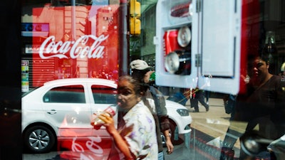 In this Oct. 9, 2014 file photo, a woman is reflected in a Coca-Cola store window display as she drinks a Coke in Mexico City. Mexicans are among the biggest soda drinkers in the world, so residents of the southern city of Ciudad Altamirano were hit hard when first Coca-Cola then Pepsi closed their distribution centers amid drug gang extortion demands.