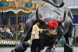 A woman pulls a 2 wheel trolley loaded with goods touches a bull statue on display outside a retail and wholesale clothing mall in Beijing, Monday, July 9, 2018. The trade war that erupted last Friday between the U.S. and China carries a major risk of escalation that could weaken investment, depress spending, unsettle financial markets and slow the global economy.