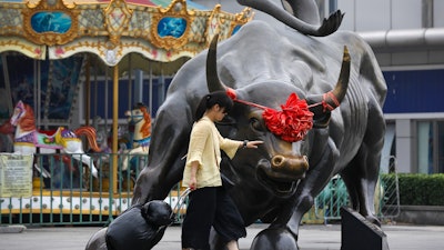 A woman pulls a 2 wheel trolley loaded with goods touches a bull statue on display outside a retail and wholesale clothing mall in Beijing, Monday, July 9, 2018. The trade war that erupted last Friday between the U.S. and China carries a major risk of escalation that could weaken investment, depress spending, unsettle financial markets and slow the global economy.