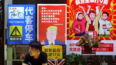 n this Monday, Aug. 13, 2018, photo, a man stands near a poster depicting a mural of U.S. President Donald Trump stating that all American customers will be charged 25 percent more than others starting from the day president Trump started the trade war against China, on display outside a restaurant in Guangzhou in south China's Guangdong province. The recent trade war between the world's two biggest economies has forced many multinational companies to reschedule purchases and rethink where they buy materials and parts to try to dodge or blunt the effects of tit-for-tat tariffs between Washington and Beijing.