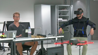 Robotics expert on the Ecole Polytechnique Federal de Lausanne, EPFL team, Matteo Macchini, right, displays the fly controls in a virtual reality scenario as he controls the flight of a drone in foreground, while observed by a fellow member of the team, July 23, 2018, in Lausanne, Switzerland. Scientists, engineers and robotics experts at the Swiss university have teamed up to develop a jacket that allows hands-free, torso-controlled flying of drones.