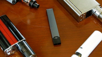 This Tuesday, April 10, 2018 file photo shows vaping devices, including a Juul, center, that were confiscated from students at a high school in Marshfield, Mass. On Tuesday, Nov. 13, 2018, San Francisco-based Juul Labs Inc. announced it had stopped filling orders for its mango, fruit, creme and cucumber pods but not menthol and mint. It will sell all flavors through its website and limit sales to those 21 and older.