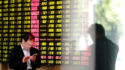 A man looks at his smartphone near a display showing stock prices at a brokerage house in Shanghai Monday, May 6, 2019. China's benchmark Shanghai Composite index dives on U.S. President Donald Trump threat of more China tariffs. At right is a reflection off a display board.