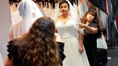 In this Friday, June 21, 2019, photo Ann Campeau,, right, owner of Strut Bridal, fits a new dress on inventory manager Stefanie Zuniga at her shop in Tempe, Ariz. Cut-rate prices on websites that sell wedding dresses direct from China put pressure on Campeau, who owns four bridal shops in California and Arizona. She has had customers come in after seeing low-end gowns online and expecting to get a dress at a similar price.