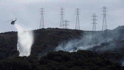 In this Oct. 10, 2019, file photo, a helicopter drops water near power lines and electrical towers while working at a fire on San Bruno Mountain near Brisbane, Calif. California’s Pacific Gas & Electric is faced regularly with a no-win choice between risking the start of a deadly wildfire or immiserating millions of paying customers by shutting off the power.