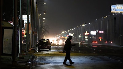 In this May 2, 2019, file photo, a pedestrian crosses Gratiot Street at Outer Drive East under a bright streetlight flanked by dimmer streetlights in Detroit. Leotek Electronics USA has agreed to pay $4 million to settle a lawsuit with Detroit over thousands of defective streetlights in Detroit, a newspaper reported. The city sued the company last spring, alleging that roughly 20,000 lights were failing.