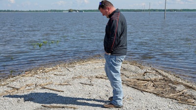 In this May 10, 2019 photo, Brett Adams stands where the road to his flooded farm disappears under flood waters, with the farm buildings visible in the background, in Peru, Neb. Adams had thousands of acres under water, about 80 percent of his land, this year. The water split open his grain bins and submerged his parents' house and other buildings when the levee protecting the farm broke.