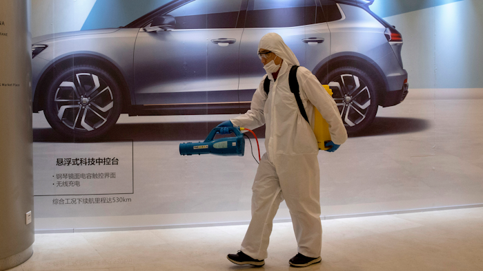 In this Feb. 12, 2020, photo, a worker disinfects a mall near an advertisement for a car in Beijing, China. Automakers are gradually reopening factories in China that were idled by anti-virus control.