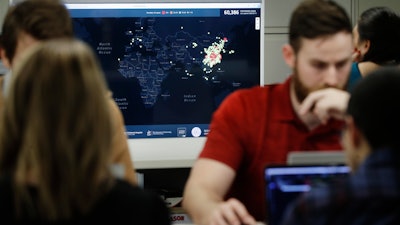 In this Thursday, Feb. 13, 2020 photo, Kyle Martin, a worker at HealthMap, a system using artificial intelligence to monitor global disease outbreaks, mines health data to keep the system up to date in a work area at Boston Children's Hospital in Boston. Early warnings of an emerging epidemic, some of them collected by HealthMap, showed AI's potential for giving global health experts a head start when bureaucratic hurdles and language barriers might get in the way. A screen behind displays a world map with colored dots marking cases of the disease known as COVID-19.