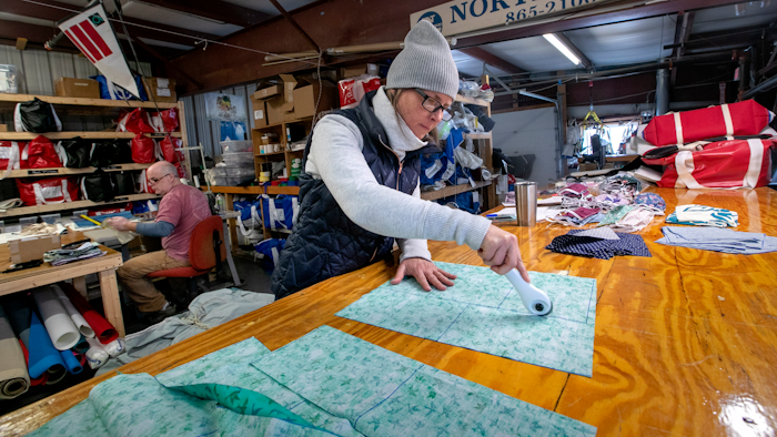 In this Monday, March 23, 2020, photo, Karen Haley cuts cotton fabric for masks to be given to caregivers during the coronavirus outbreak, at the North Sails shop in Freeport, Maine. The sail-maintenance business has converted part of its operation towards stitching masks instead of sails. Owner Eric Baldwin stitches masks in background.