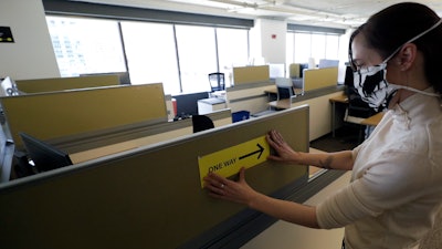 This Thursday, May 7 photo shows Interior Designer Stephanie Jones at the design firm Bergmeyer putting up a safe distancing reminder indicating one way foot traffic to a cubicle at the firms offices in Boston.