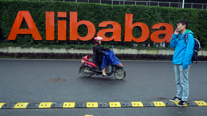 In this May 27, 2016, file photo, a man talks on his phone as a woman rides on an electric bike past a company logo at the Alibaba Group headquarters in Hangzhou in eastern China's Zhejiang province.