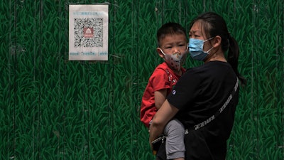 A woman wearing a face mask carries a masked child stands against a board displaying a health QR code on Wednesday, May 18, 2022, in Beijing. Angry bank customers who traveled to a city in central China attempting to retrieve their savings from troubled rural banks were stopped in their tracks by a common technology: a QR code.
