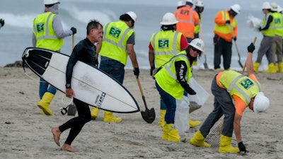 A surfer walks by as workers in protective suits continue to clean the contaminated beach in Huntington Beach, Calif., on Oct. 11, 2021.