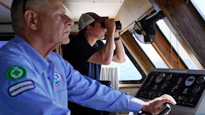 Aaron Smith, President and CEO of the Offshore Marine Service Association, center, peers through binoculars at ships installing the South Fork Wind project, as Capt. Rick Spaid, left, pilots the vessel Jones Act Enforcer, Tuesday, July 11, 2023, off the coast of Rhode Island.
