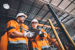 Two construction workers in orange safety vests review tablet in industrial warehouse