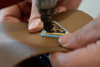 Close-up of hands attaching a gold Prada logo tag to tan leather material