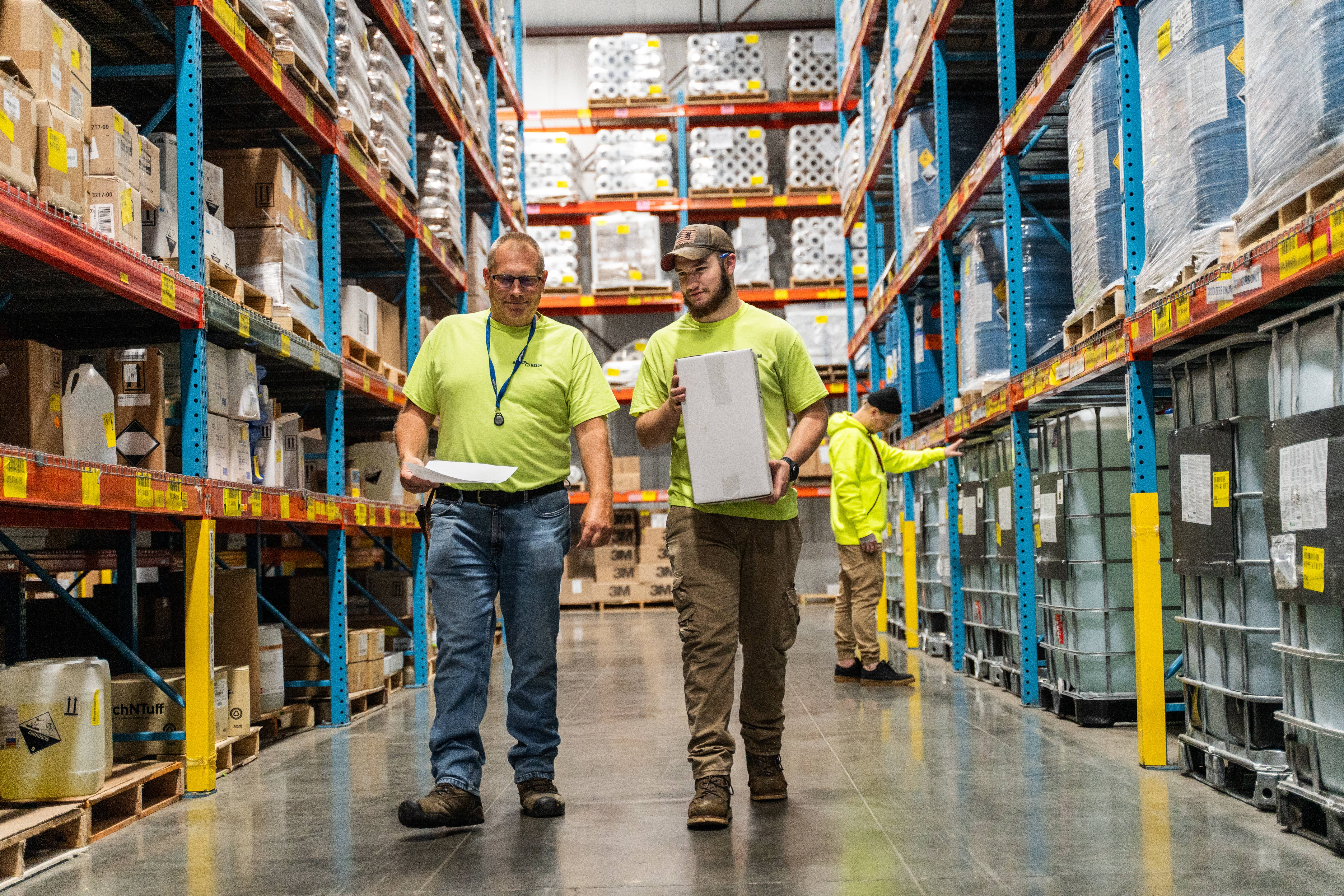 Two warehouse workers in safety vests walking through industrial shelving with inventory and products