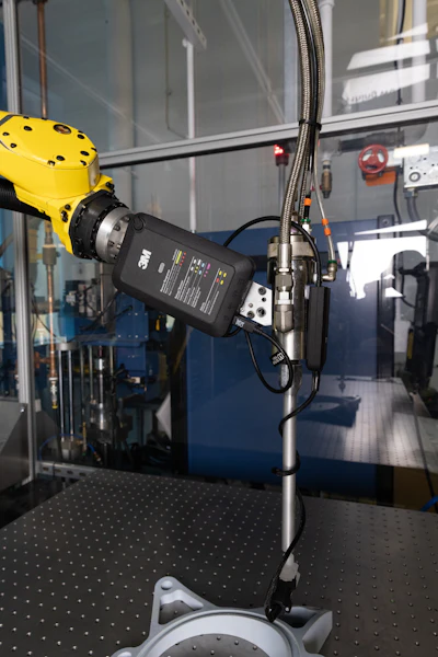 Yellow robotic arm with 3M power adapter mounted on laboratory equipment frame with metal cables and control panel