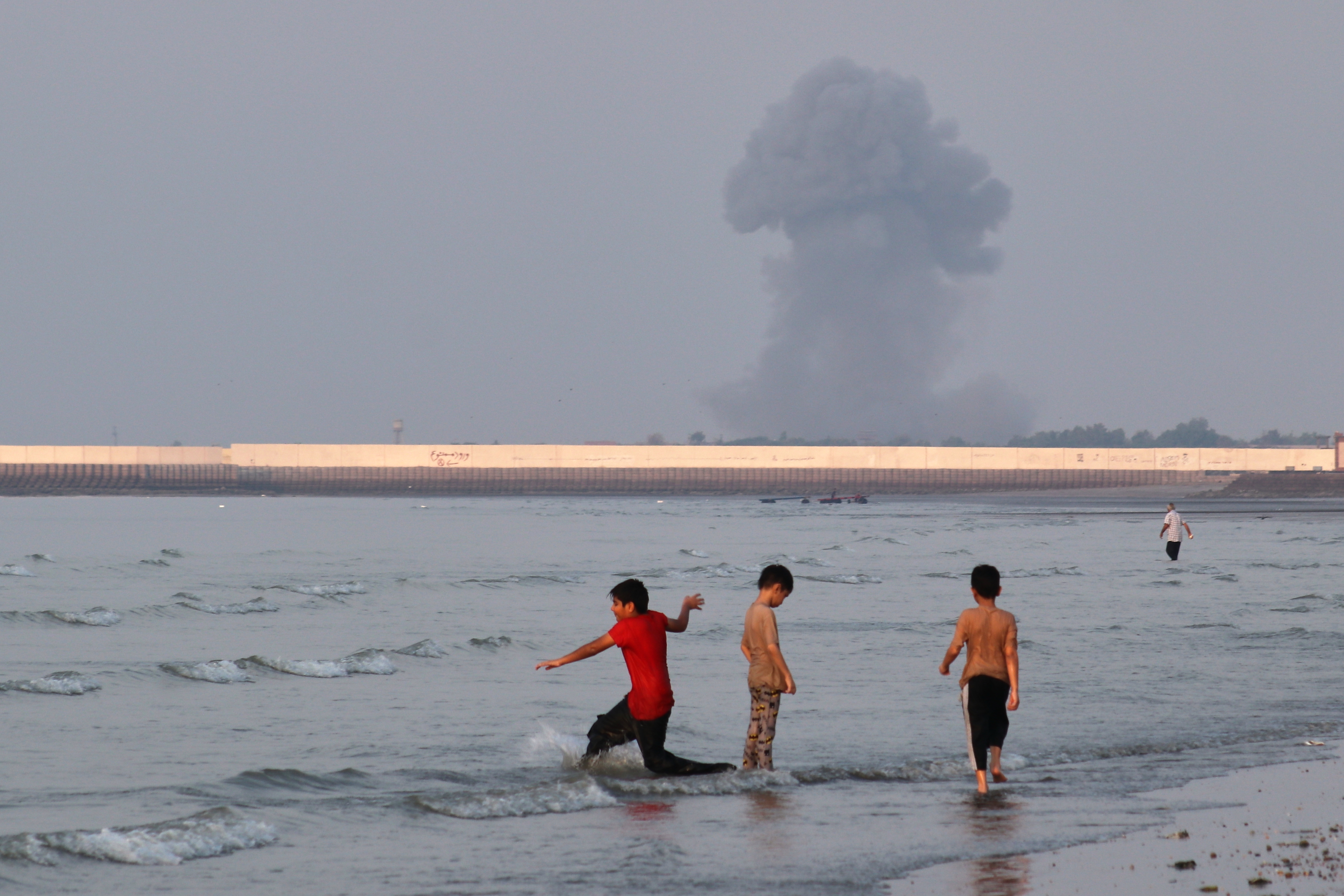 Three boys play in the shallow waters of the Strait of Hormuz as a plume of smoke rises from an explosion in the background off Bandar Abbas, Iran, July 13, 2026.