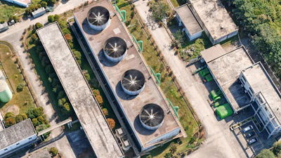 Aerial view of industrial facility with four circular cooling towers on rooftop surrounded by landscaped grounds and adjacent buildings