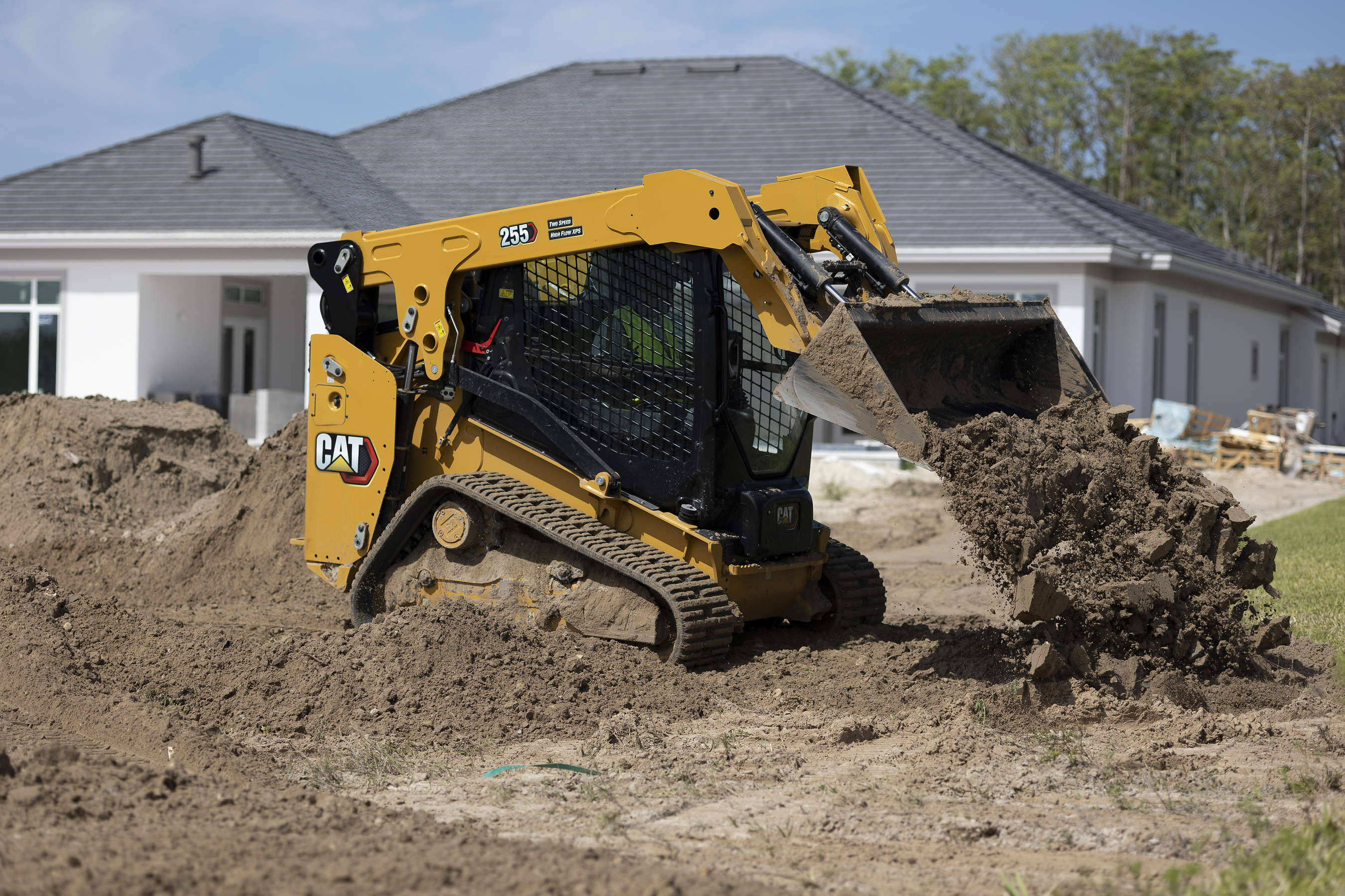 Yellow CAT 259 compact track loader moving soil at residential construction site with white house in background