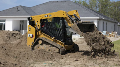 Yellow CAT 259 compact track loader moving soil at residential construction site with white house in background