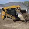 Yellow CAT 259 compact track loader moving soil at residential construction site with white house in background
