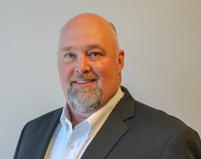 Professional headshot of a smiling man with gray beard wearing a dark blazer and light blue shirt
