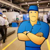 Cartoon mechanic in blue uniform with sad expression stands with crossed arms in industrial manufacturing facility