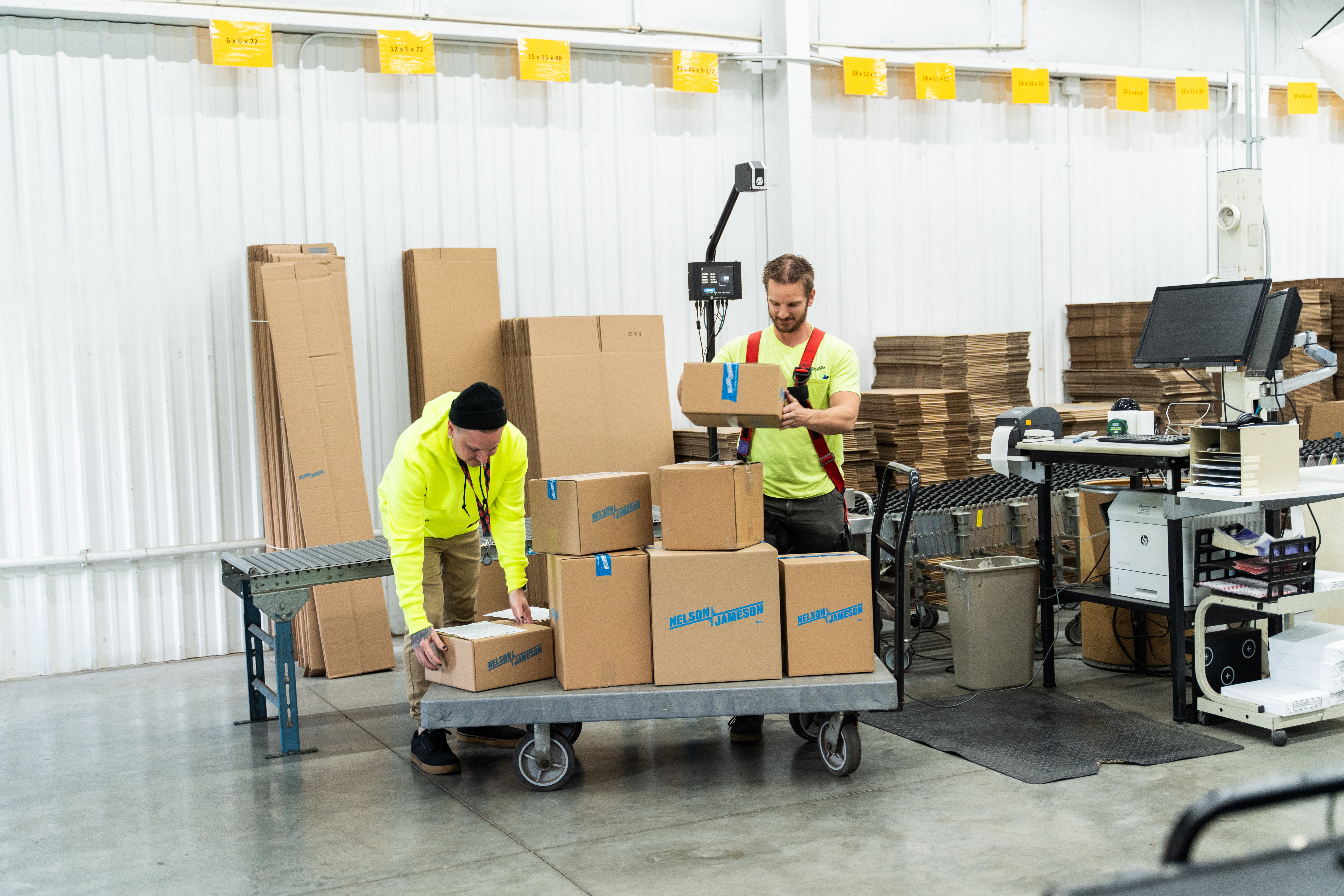 Two warehouse workers in safety vests processing cardboard boxes on a cart in a fulfillment center