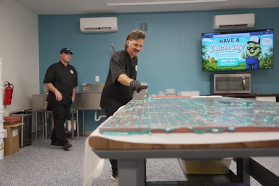 Two staff members demonstrate cornhole in a recreational room with turquoise walls and air conditioning units.