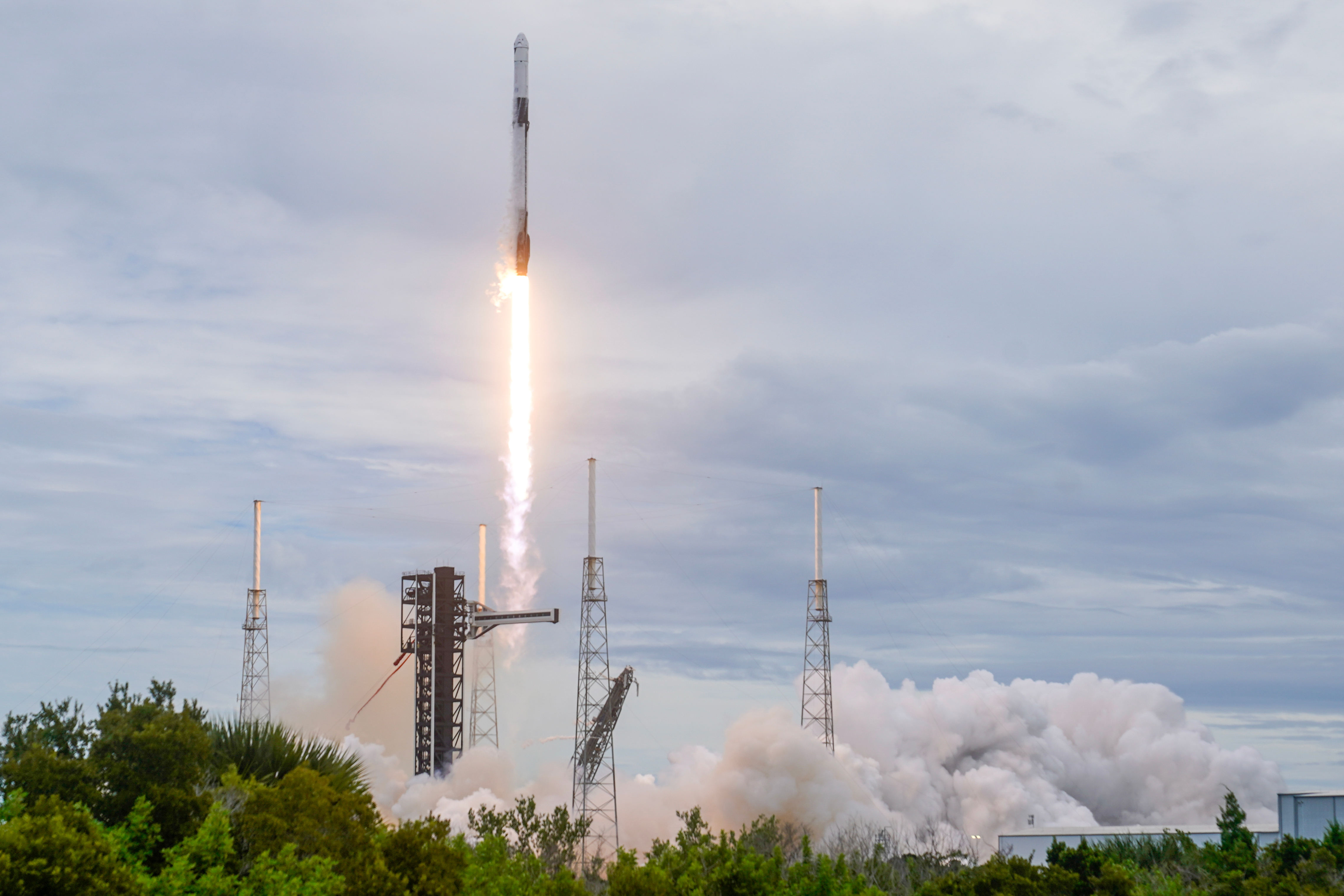SpaceX Falcon 9 rocket launches from coastal launch pad with bright engine flames and white smoke clouds against cloudy sky