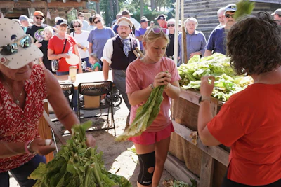 Community members examine fresh lettuce at an outdoor farmers market or harvest event with crowds gathered around.