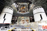 Two white spacecraft modules with black checkered docking targets in assembly facility with metal framework and equipment overhead.