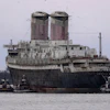 Large cargo ship assisted by tugboats in port waters with industrial twin smokestacks visible