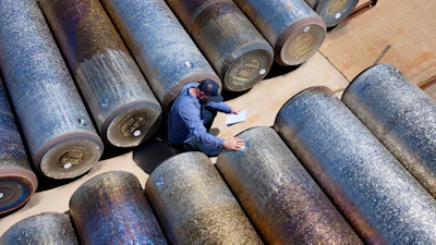 Worker in blue surveying large industrial metal pipes arranged on sandy ground from above