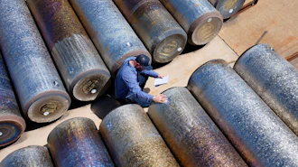 Worker in blue surveying large industrial metal pipes arranged on sandy ground from above