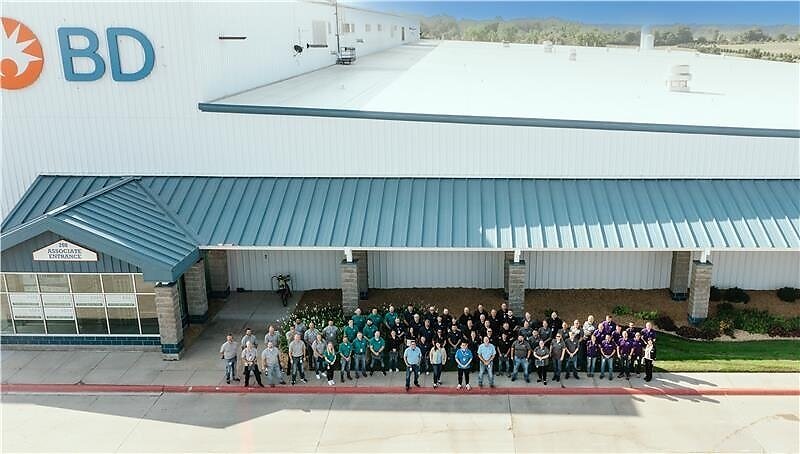 Large group of BD company employees standing together outside a modern industrial facility with white and teal metal roofing.