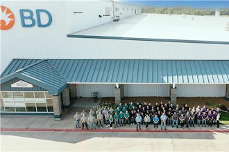 Large group of BD company employees standing together outside a modern industrial facility with white and teal metal roofing.