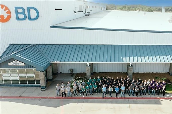 Large group of BD company employees standing together outside a modern industrial facility with white and teal metal roofing.